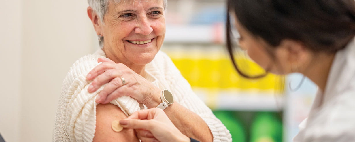 An elderly woman smiles as a healthcare professional administers a vaccination on her upper arm