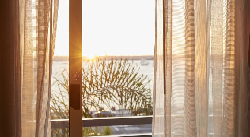 Close up of some brown curtains with morning sun shining through. In the background you can see a tree and some water
