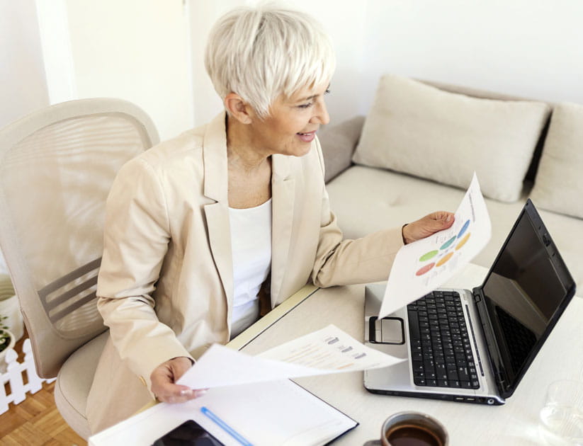 Mature businesswoman sitting in home office and using her laptop while looking at her finances and paperwork