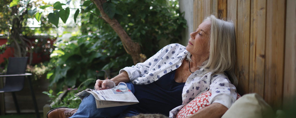 A mature woman reading a newspaper taking a nap on outdoor seating in a garden