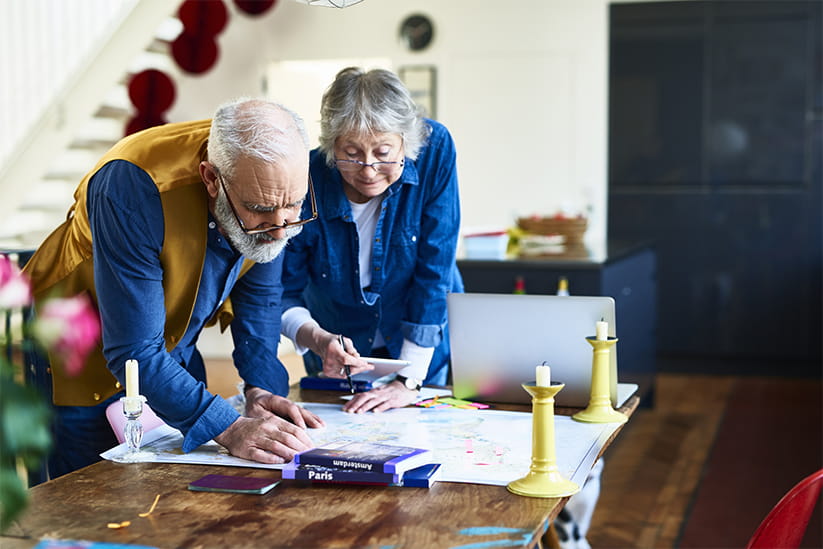 Retired couple looking at map and organising route, guide books on table, using sticky notes to highlight tourist destinations and landmarks