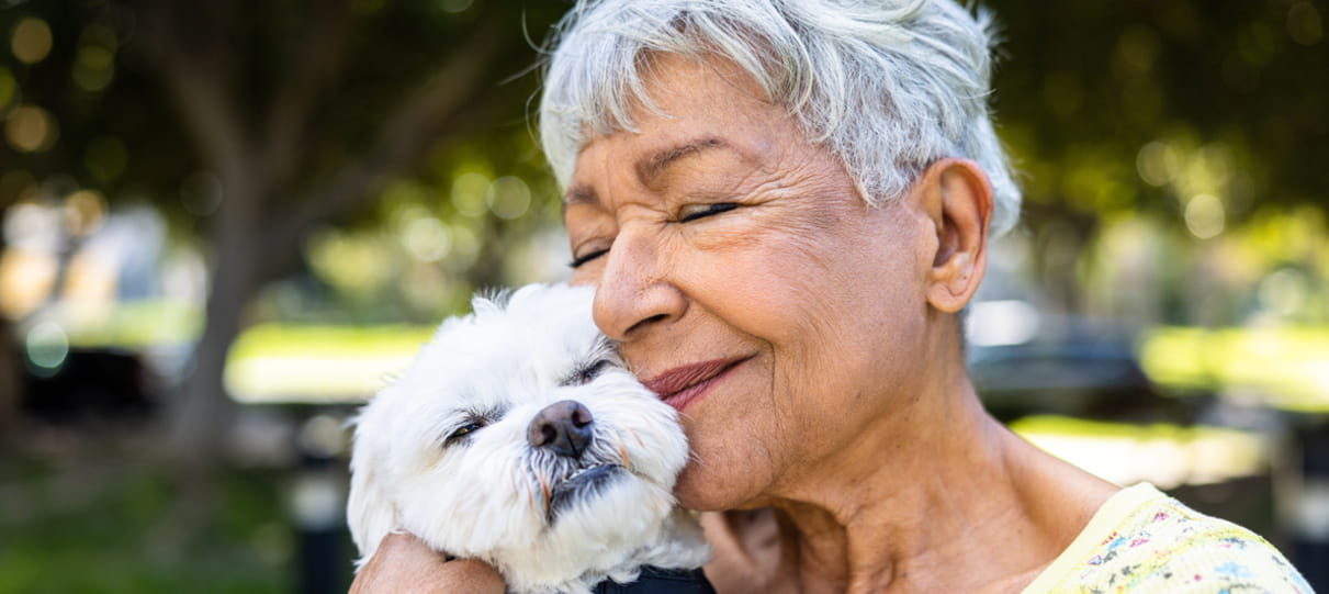 An older woman with short grey hair snuggles up outdoors with her white fluffy dog