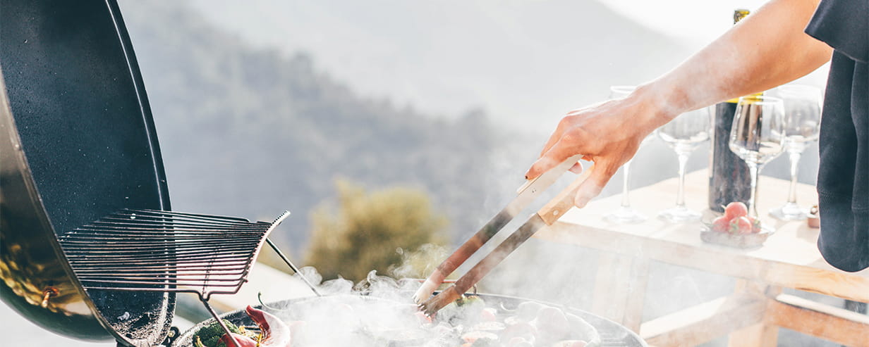 An older person tending a barbecue with a bottle of wine and wine glasses on a table in the background