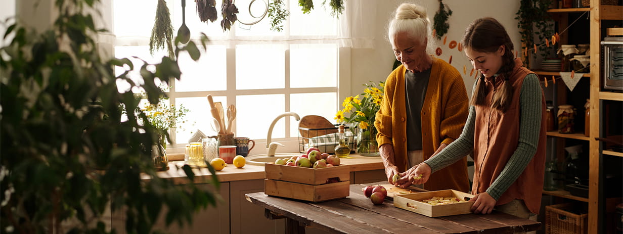 Younthful girl with pigtails putting slices apples on wooden tray while standing next to grandmother