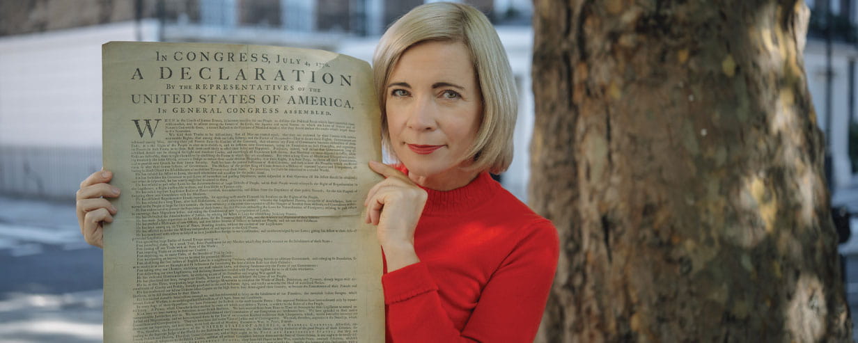 Lucy Worsley in a red jumper holding up the American Declaration of Independence