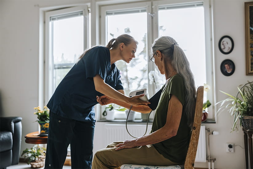Female caregiver examining blood pressure of senior woman sitting on chair at nursing home