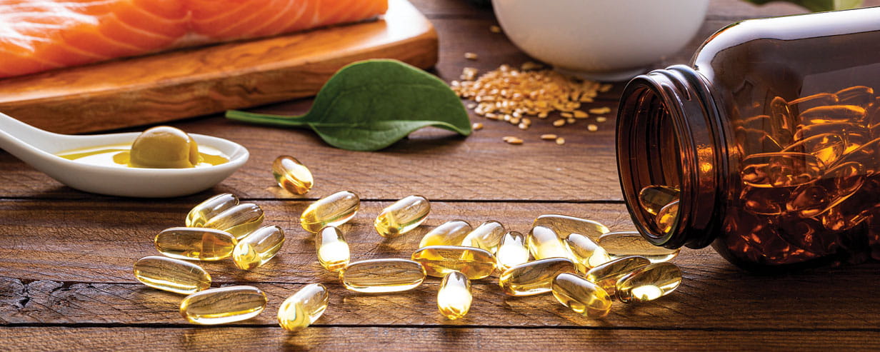 Front view of many fish oil capsules spilling out from the bottle surrounded by an assortment of food rich in omega-3 such as salmon, flax seeds, broccoli, sardines, spinach, olives and olive oil.