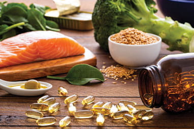 Front view of many fish oil capsules spilling out from the bottle surrounded by an assortment of food rich in omega-3 such as salmon, flax seeds, broccoli, sardines, spinach, olives and olive oil.