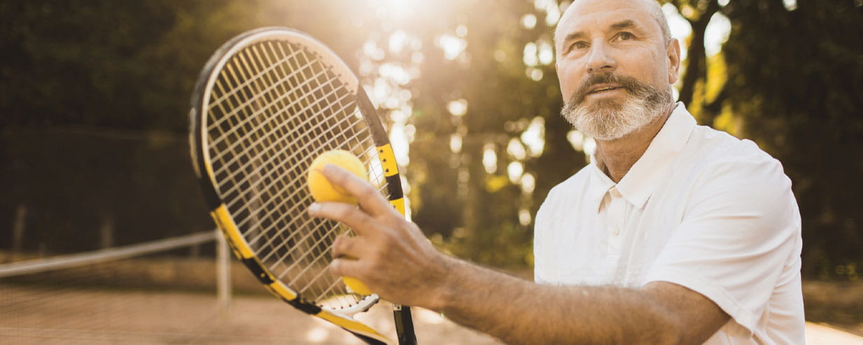Active senior man with beard with tennis racket before serve with the sunlight streaming in from behind him
