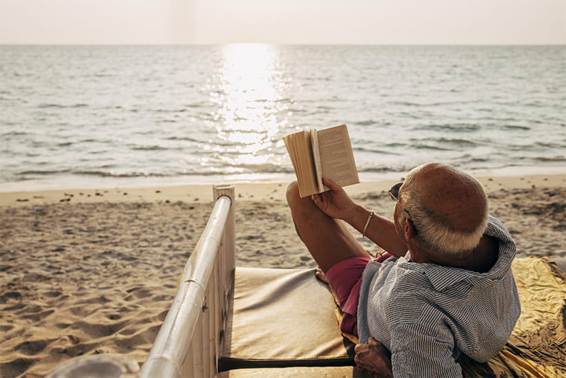 Relaxed senior man lying on bed while reading book on beach at sunset