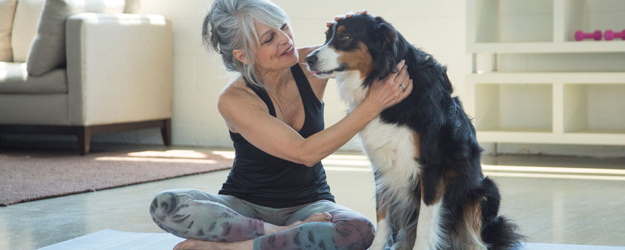 Mature woman sitting cross legged on a yoga mat petting a big fluffy dog sitting next to her