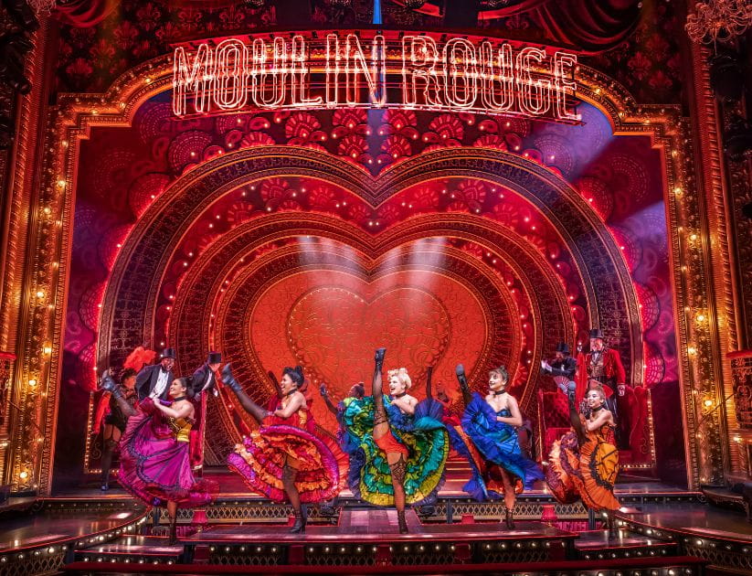 Women dancing on stage in colourful dresses below a 'Moulin Rouge' sign