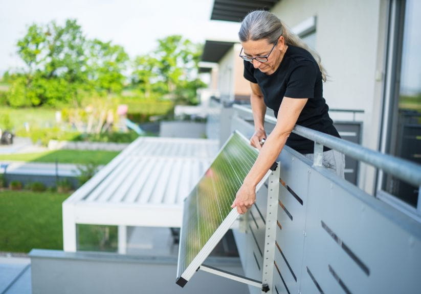 A woman leans over her balcony to attach a plug in solar panel to her balcony