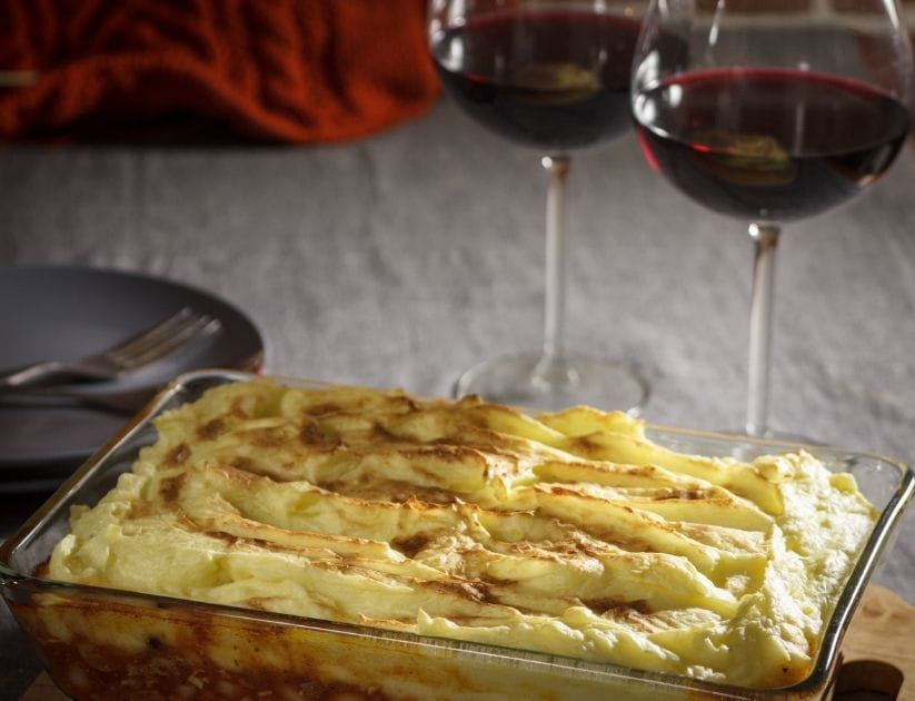 A Pyrex dish with shepherds pie and two glasses of red wine next to it