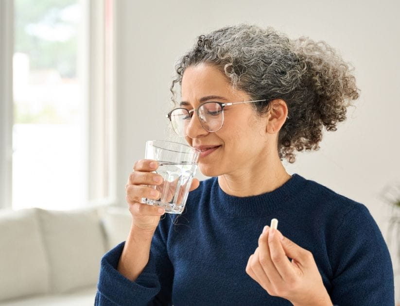 Woman of middle age taking vitamin pill while holding glass of water at home