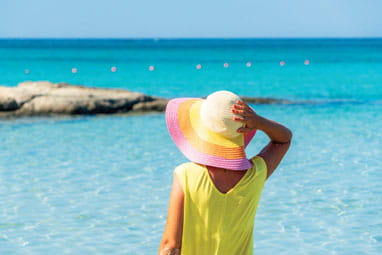 A woman in a yellow t-shirt and striped sun hat facing out towards an azure blue sea