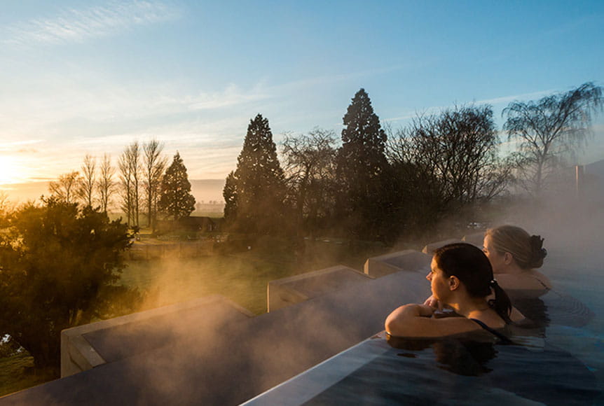 Two people in an infinity pool looking out over a garden at sunset