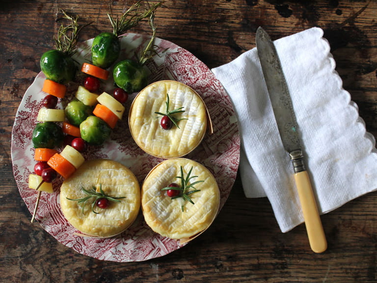 Three baked Camembert cheeses next to three vegetable skewers on a plate