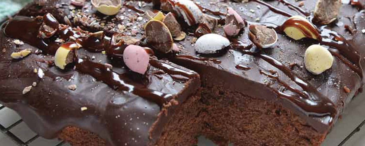 An Easter egg brownie cake, topped with mini Easter eggs, on a cooling rack with one square removed