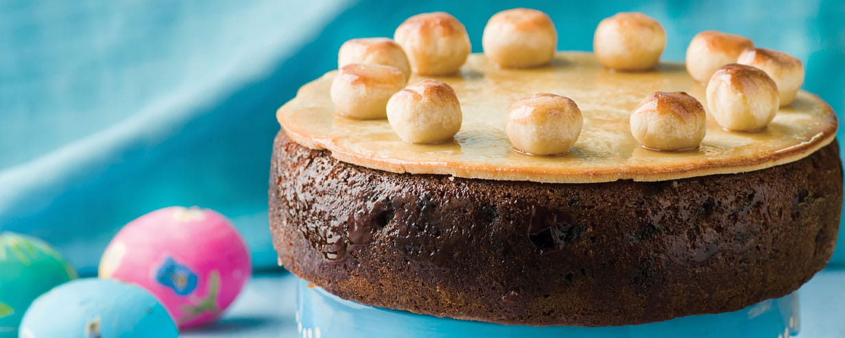 A traditional Easter Simnel cake on a stand with a blue frill with painted Easter eggs on the right