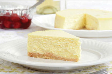 A slice of New York cheese cake with the rest of the cake in the background and a bowl of berries.