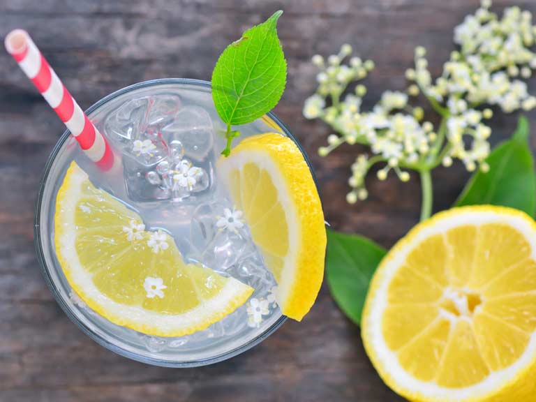 a glass topped with two slices of lemon, a white and red striped straw next to elderflowers and half a lemon