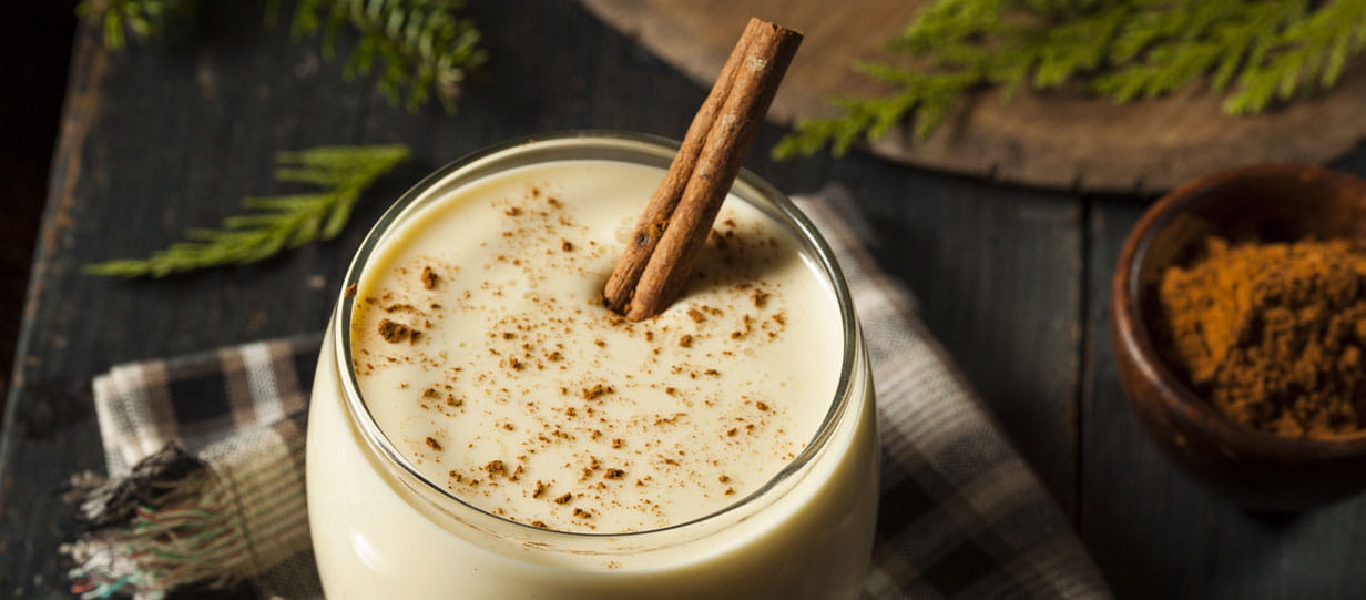 A close-up of a glass of eggnog with a stick of cinnamon and a dusting of nutmeg