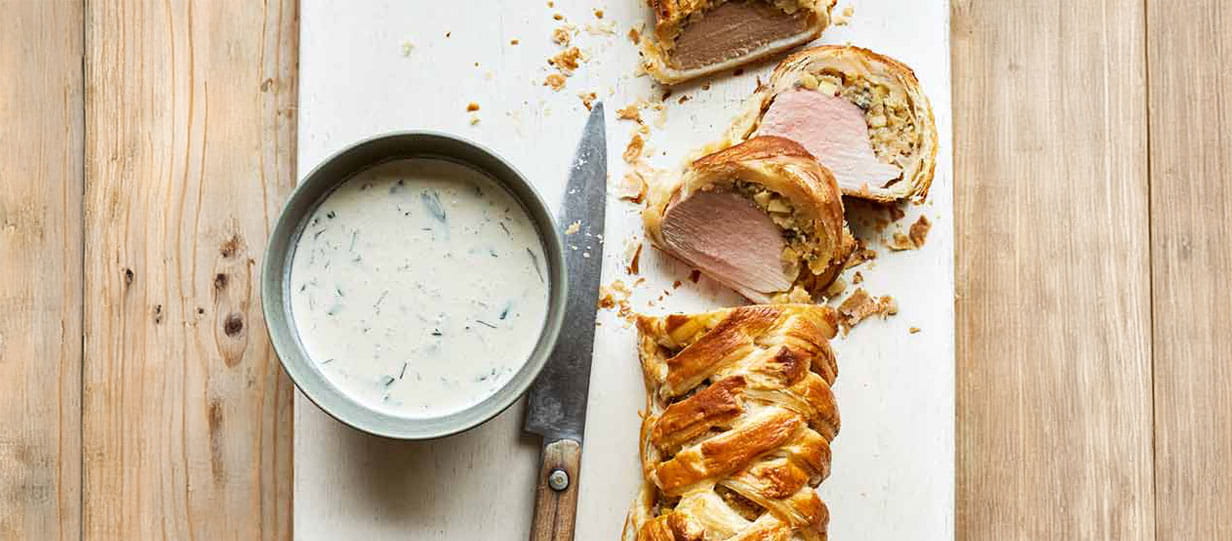 A plaited pastry with roasted pork inside and a dish of white sauce next to it