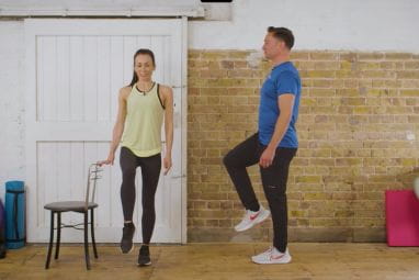 A woman and man do balance exercises in a room with a chair