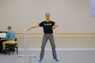 A mature ballet dancer holds a pose in a ballet studio with a piano playing behind him