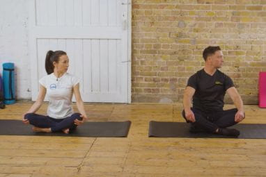 A man and woman are sat cross legged on the floor twisting their necks to one side to demonstrate a stretch