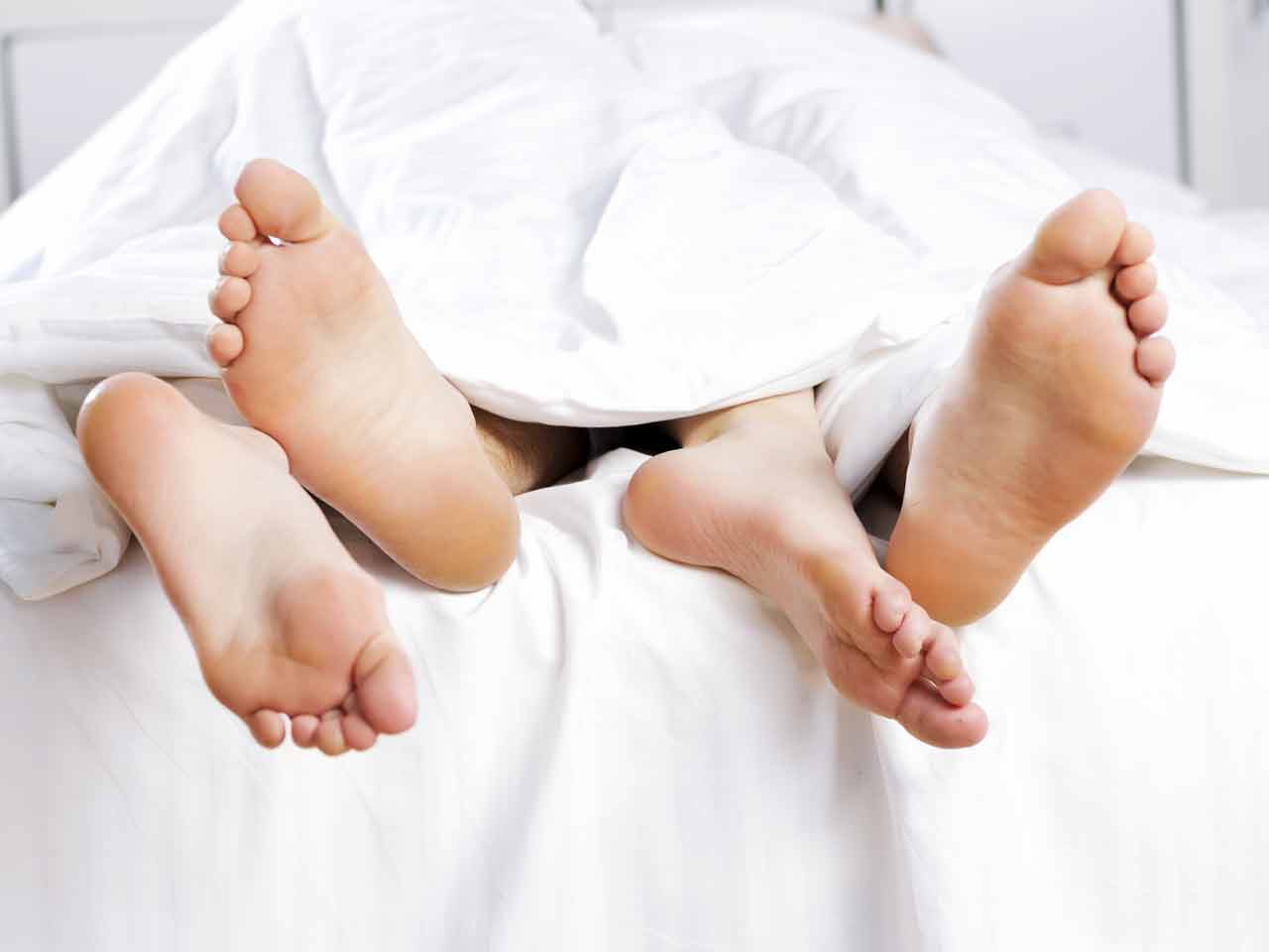 Two pairs of feet intertwined poking out the bottom of a duvet on a bed