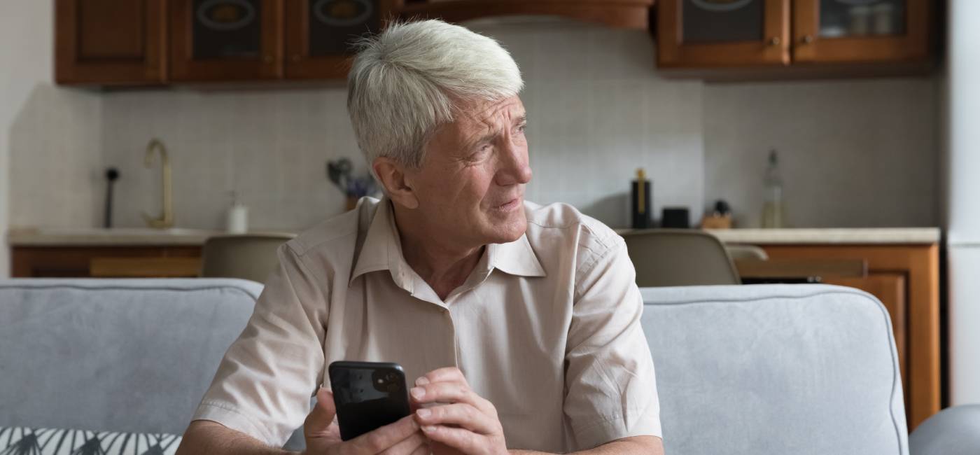 A mature male sitting in a sofa looking out the window looking worried holding his phone