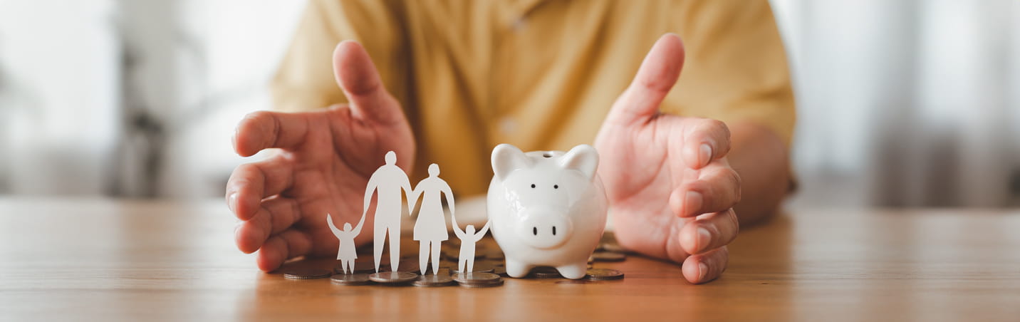 A pair of hands open on a table with a paper cutout family and a piggy bank between them