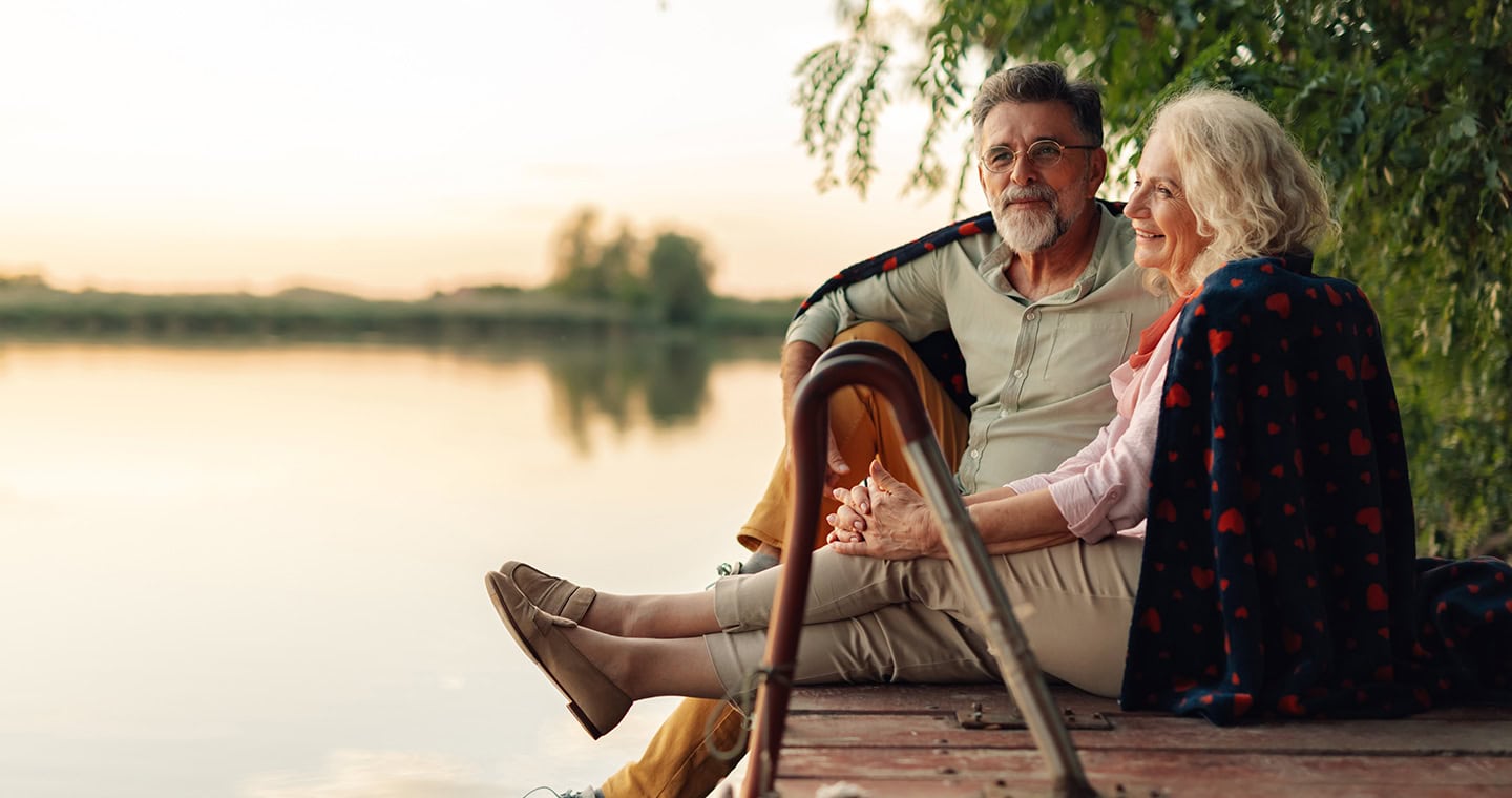 Mature couple relaxing on a pier, sharing a blanket and enjoying the sunset over the lake.