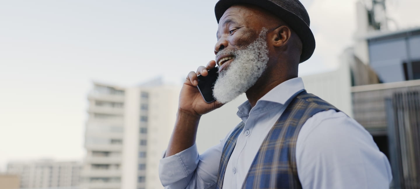 A man with a hat and beard talking outdoors, laughing and chatting on his mobile phone