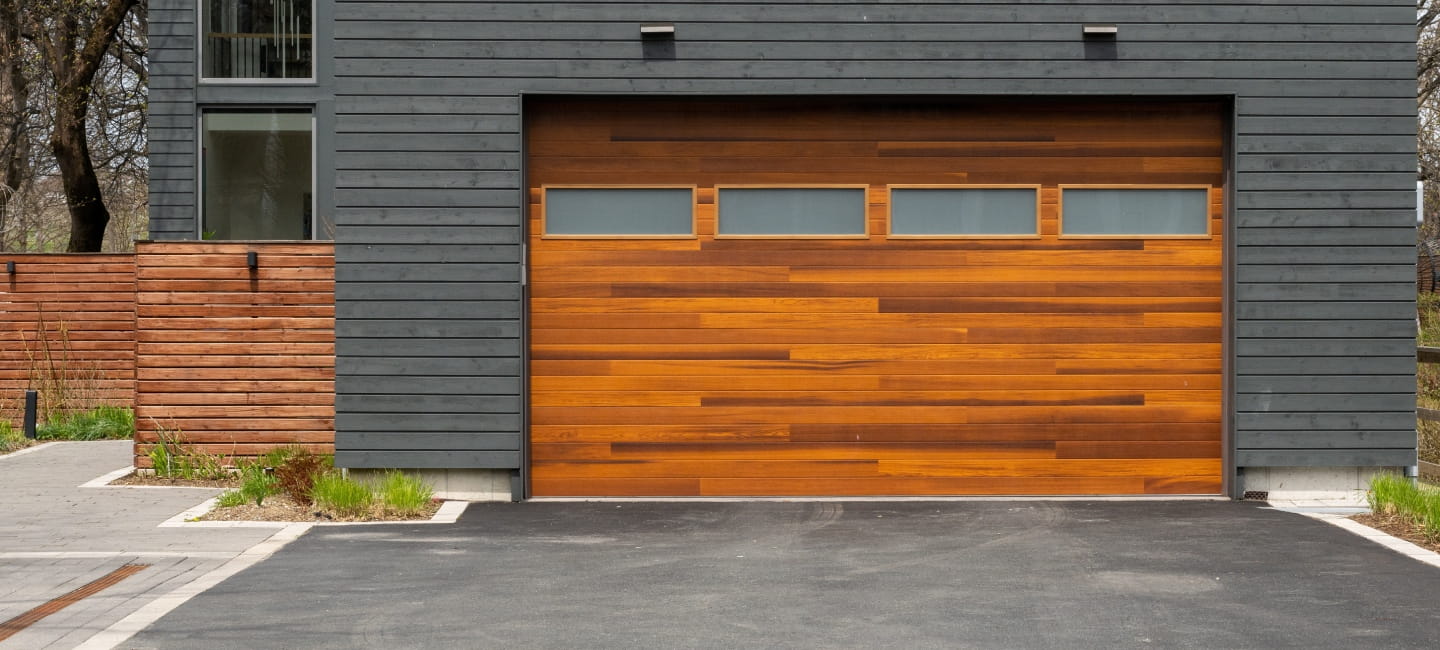A modern brown faux wooden exterior garage door with four small horizontal glass windows. The modern door is on a luxury dark grey contemporary house with a concrete driveway. 