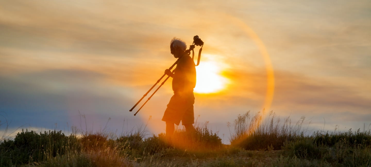 A seasoned photographer with grey hair blowing in the wind, walks at sunset with his tripod resting on his shoulder. He is in partial silhouette with the sun directly behind him.