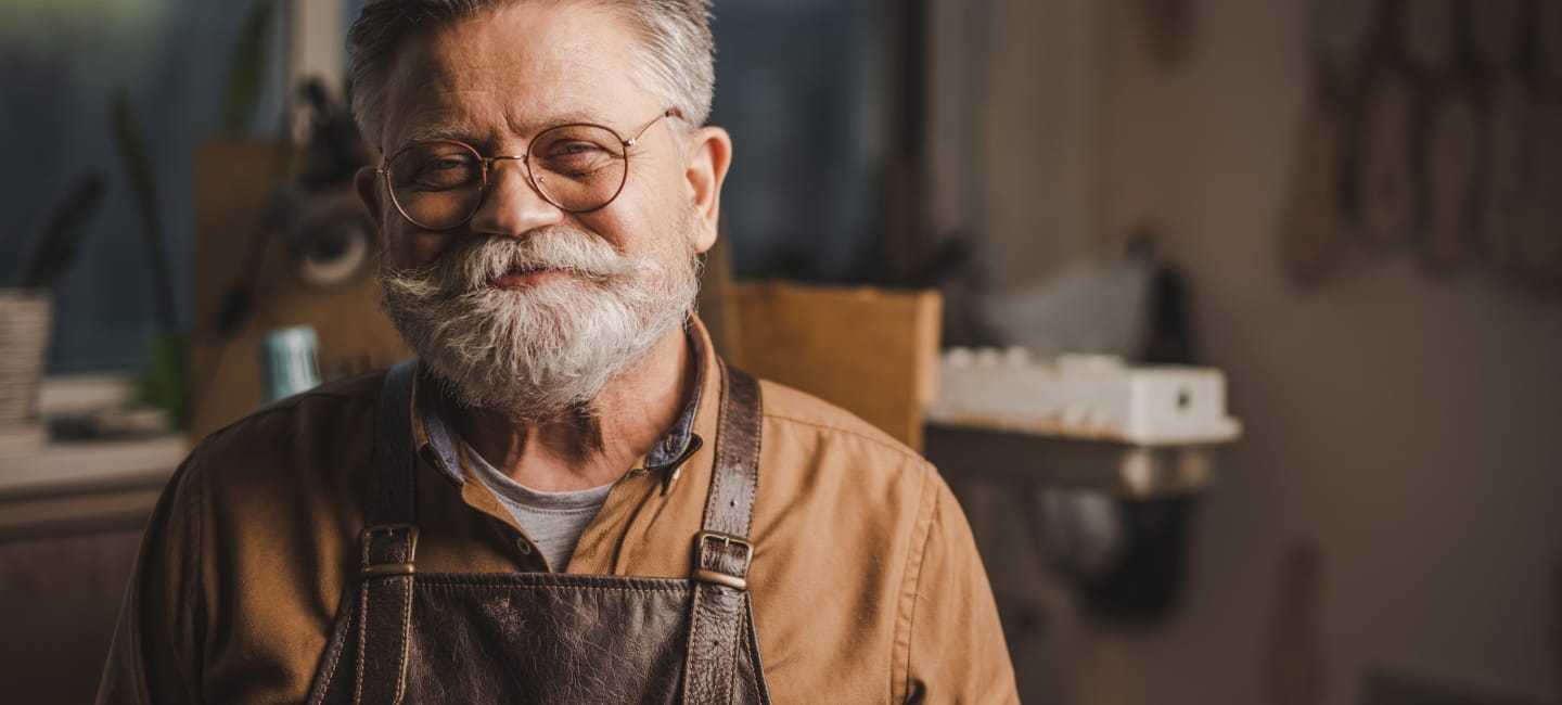 Happy, senior shoemaker in glasses smiling at camera while standing in workshop
