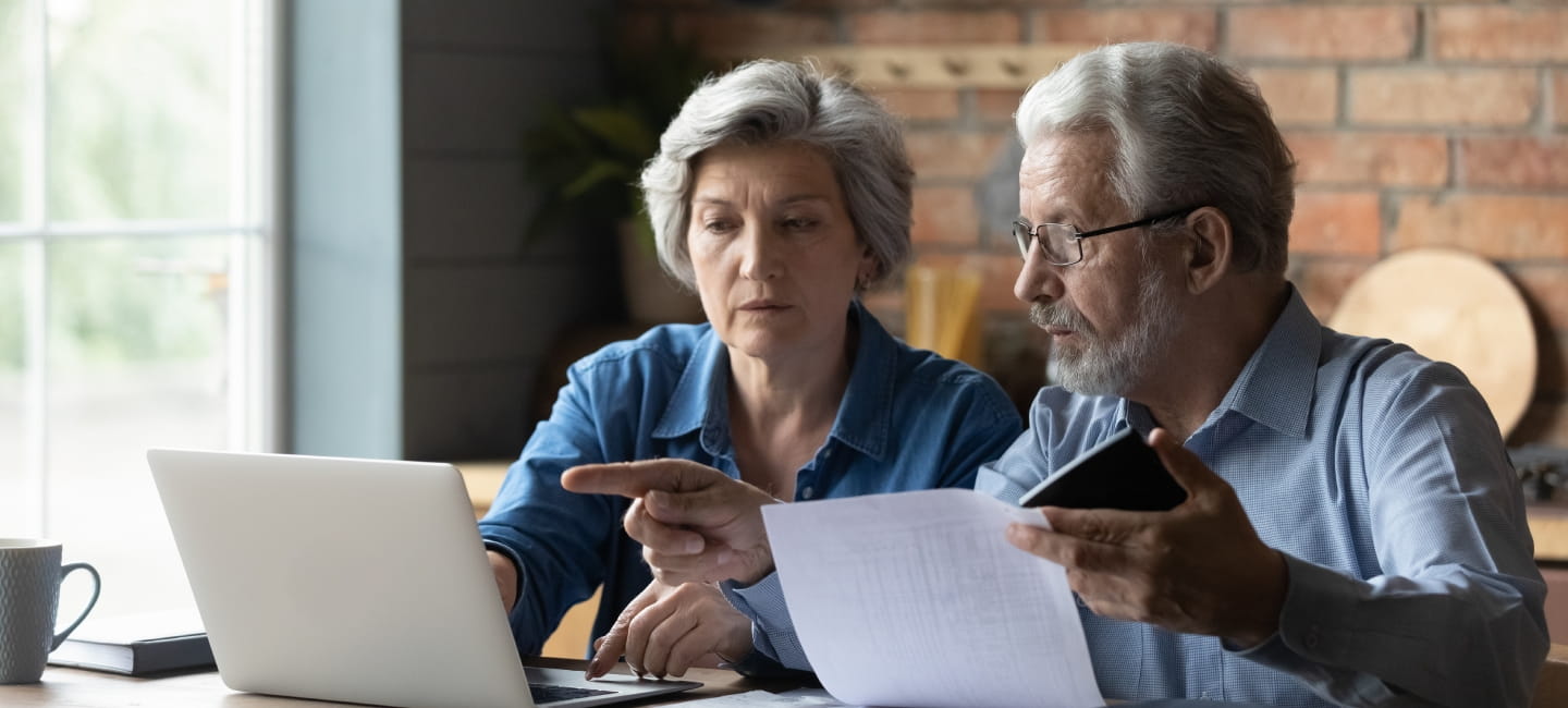 A man and woman sit at a table with a laptop. The woman holds a paper, while the main points at the laptop screen, with focused. 