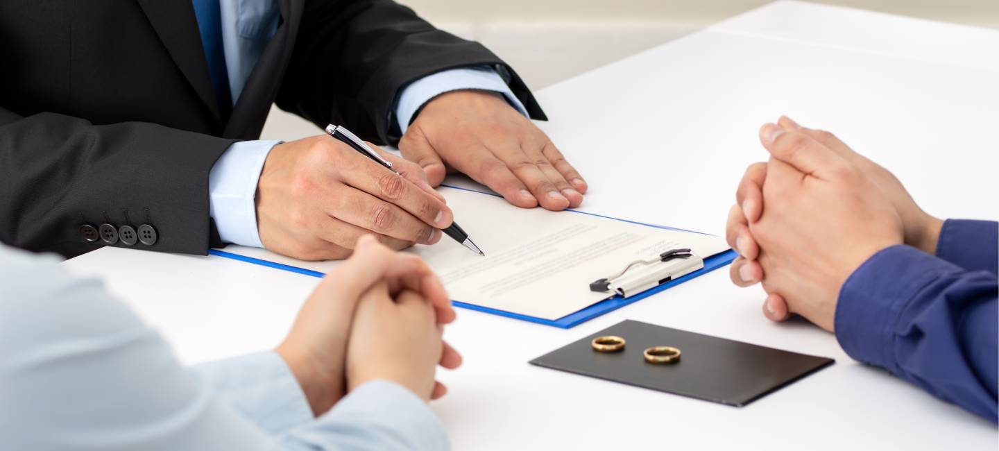 A couple are shown with their hands on a table, talking to a person in a dark suit who is filling in a document