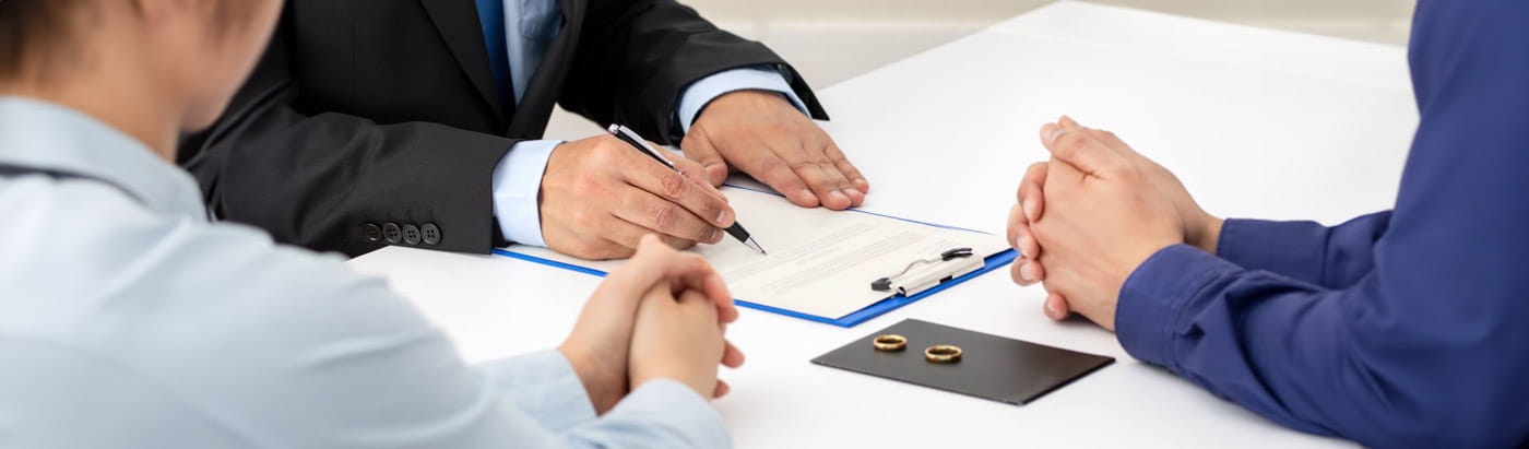 A couple are shown with their hands on a table, talking to a person in a dark suit who is filling in a document