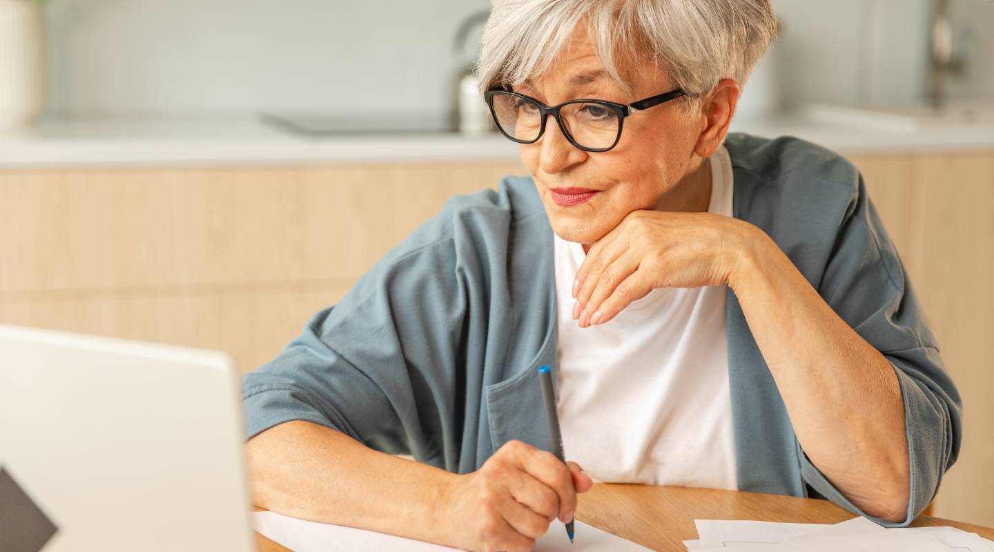 Middle aged senior woman sit with laptop and paper document, writing down pension payments