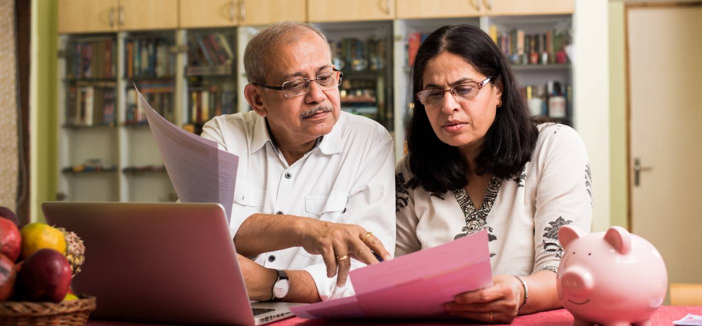 Senior Indian/asian couple accounting, doing home finance and checking bills with laptop, calculator and money while sitting on sofa/couch or dining table at home