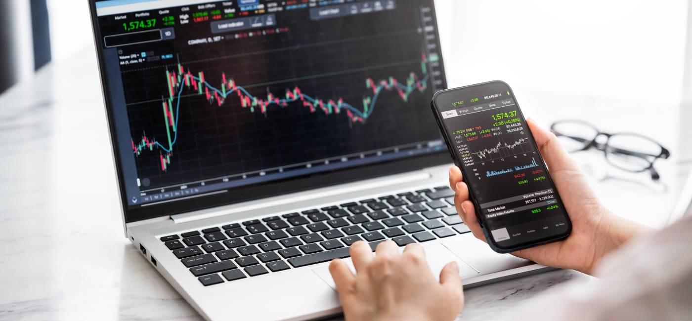 Hand holding smartphone with stock market data and using laptop display graph and chart for analyse and check before trading stocks online