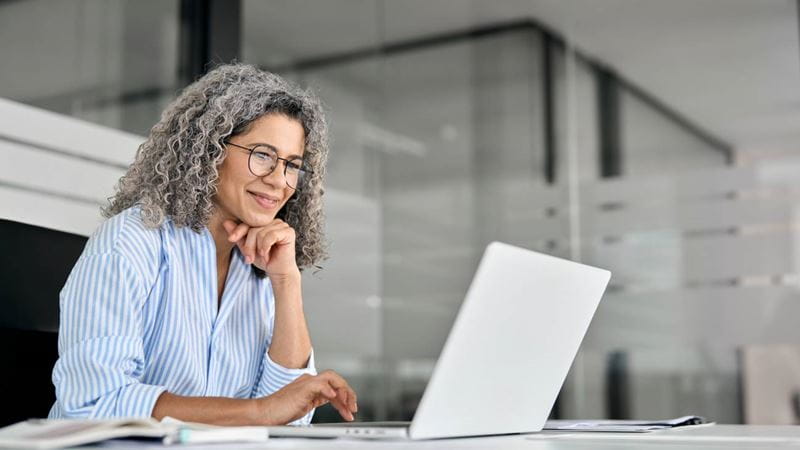 Happy older mature business woman entrepreneur in office using laptop at work, smiling professional senior middle aged female executive wearing glasses working on computer at office workplace desk.