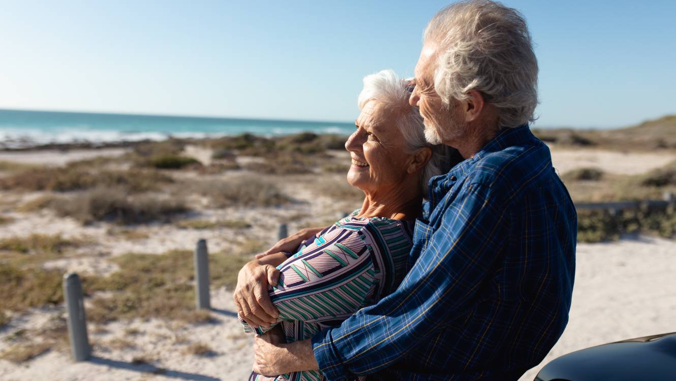 Side view close up of a senior Caucasian couple at the beach in the sun, standing and embracing, leaning against their car, smiling and admiring the view