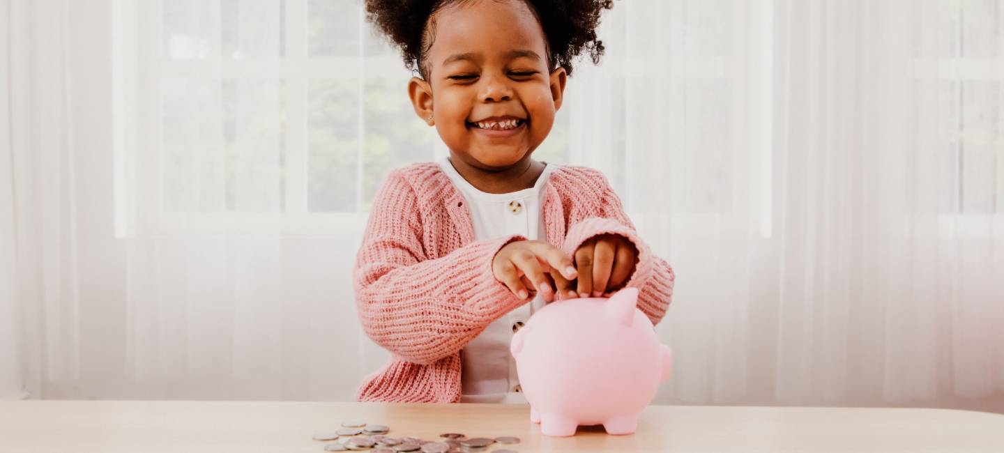 Little girl is happy and smiling as she puts her coins in a pink piggy bank sitting on a white wooden table in her home