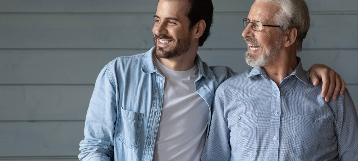 Happy two generations of men hugging, looking in distance, hugging, standing on grey wooden wall background, smiling senior father in glasses and grownup son dreaming, family unity and bonding