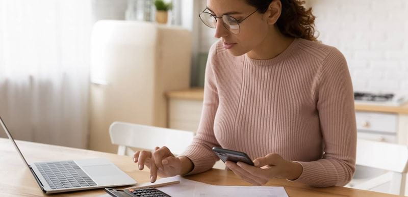 Concentrated millennial woman in eyeglasses managing monthly budget, calculating medical insurance, planning investments, paying utility household bills or taxes online using mobile phone application.